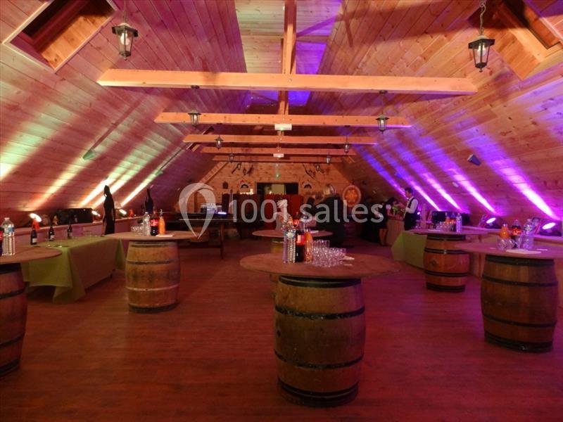 Salle en bois avec plafond en pente, éclairage coloré, tables en tonneaux et buffets alignés sur les côtés.