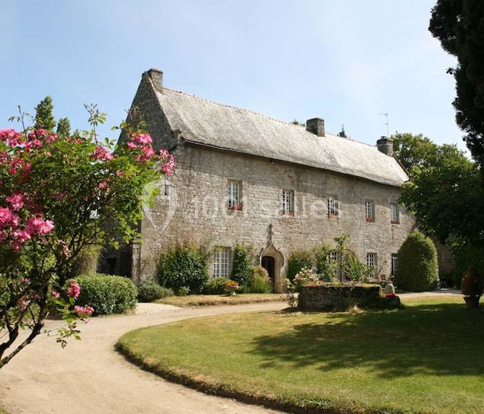 Grande maison en pierre avec toit en ardoise, entourée d'un jardin arboré et fleuri sous un ciel dégagé.