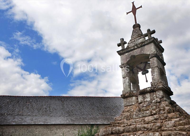 Clocher en pierre avec une croix au sommet, sur fond de ciel partiellement nuageux et toit en ardoise.