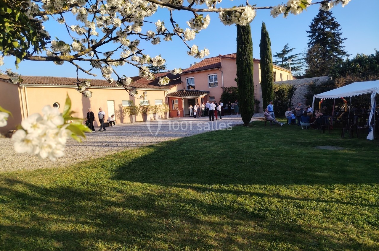 Vue d'une maison avec un jardin, des arbres en fleurs au premier plan et des personnes rassemblées près d'une tente.