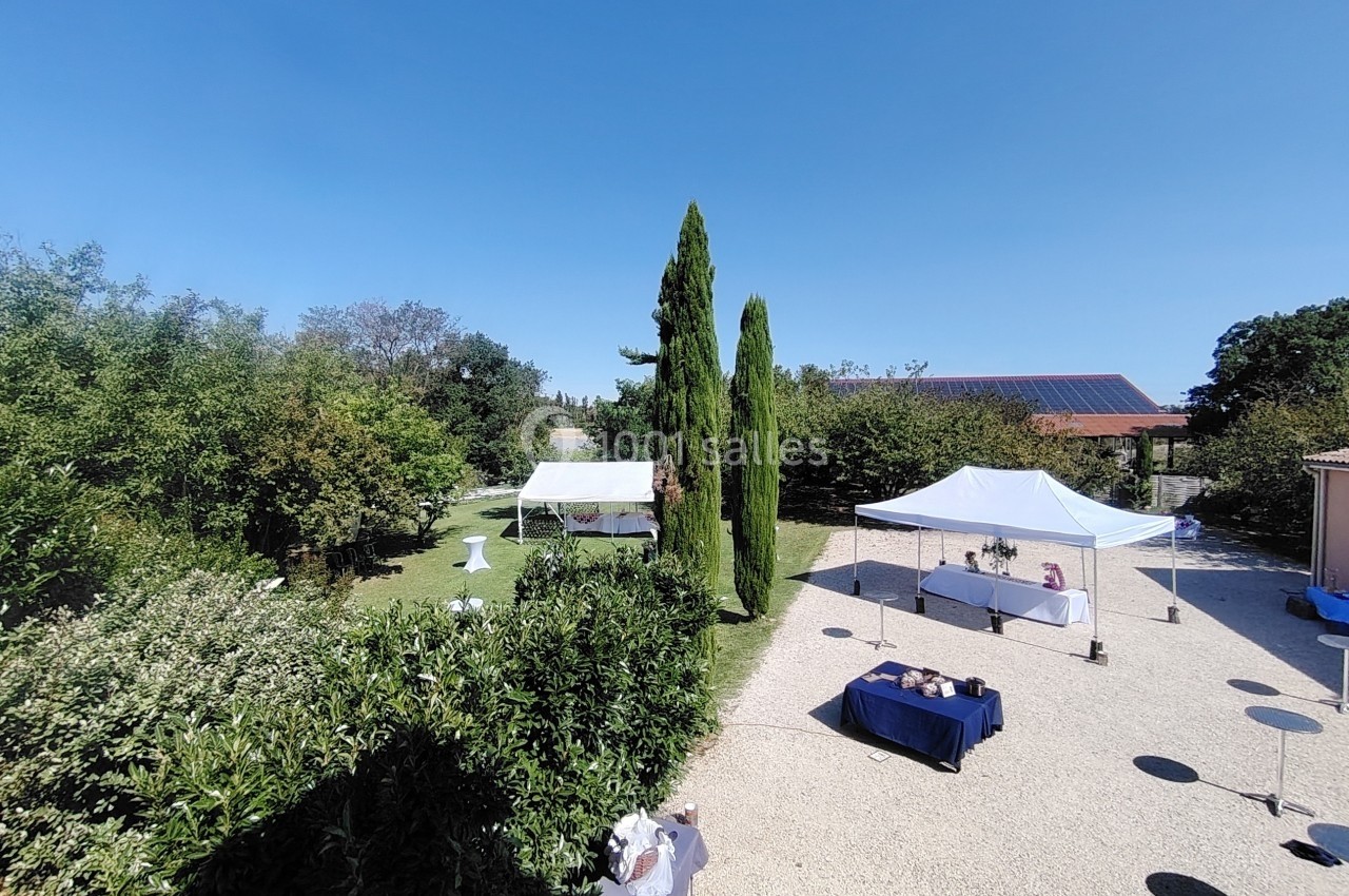 Jardin avec des arbres, des tentes blanches et des tables dressées sur une terrasse en gravier sous un ciel dégagé.