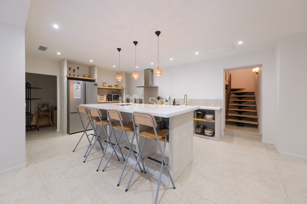Cuisine moderne avec un îlot central, tabourets hauts, éclairage suspendu et escalier visible en arrière-plan.