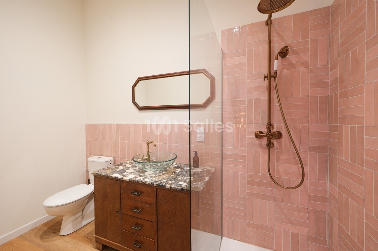 Salle de bain avec douche à l'italienne, carrelage rose, meuble en bois avec vasque en verre et miroir rectangulaire.