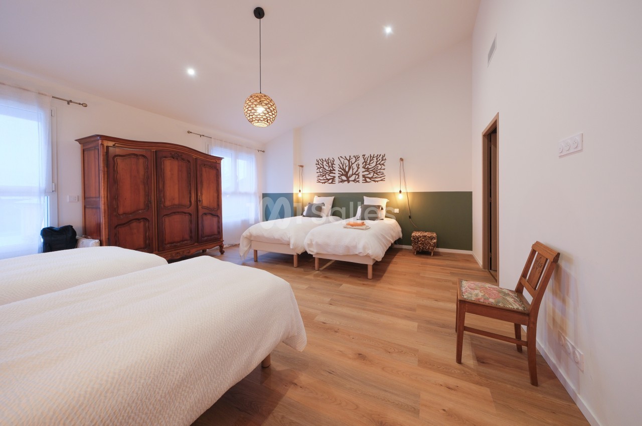 Chambre lumineuse avec deux lits simples, armoire en bois, chaise, et décoration murale sur un parquet clair.