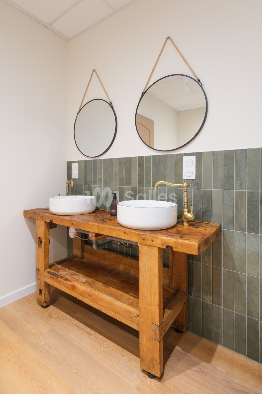 Meuble en bois avec deux vasques blanches, robinets dorés et miroirs ronds suspendus, devant un mur carrelé vert.