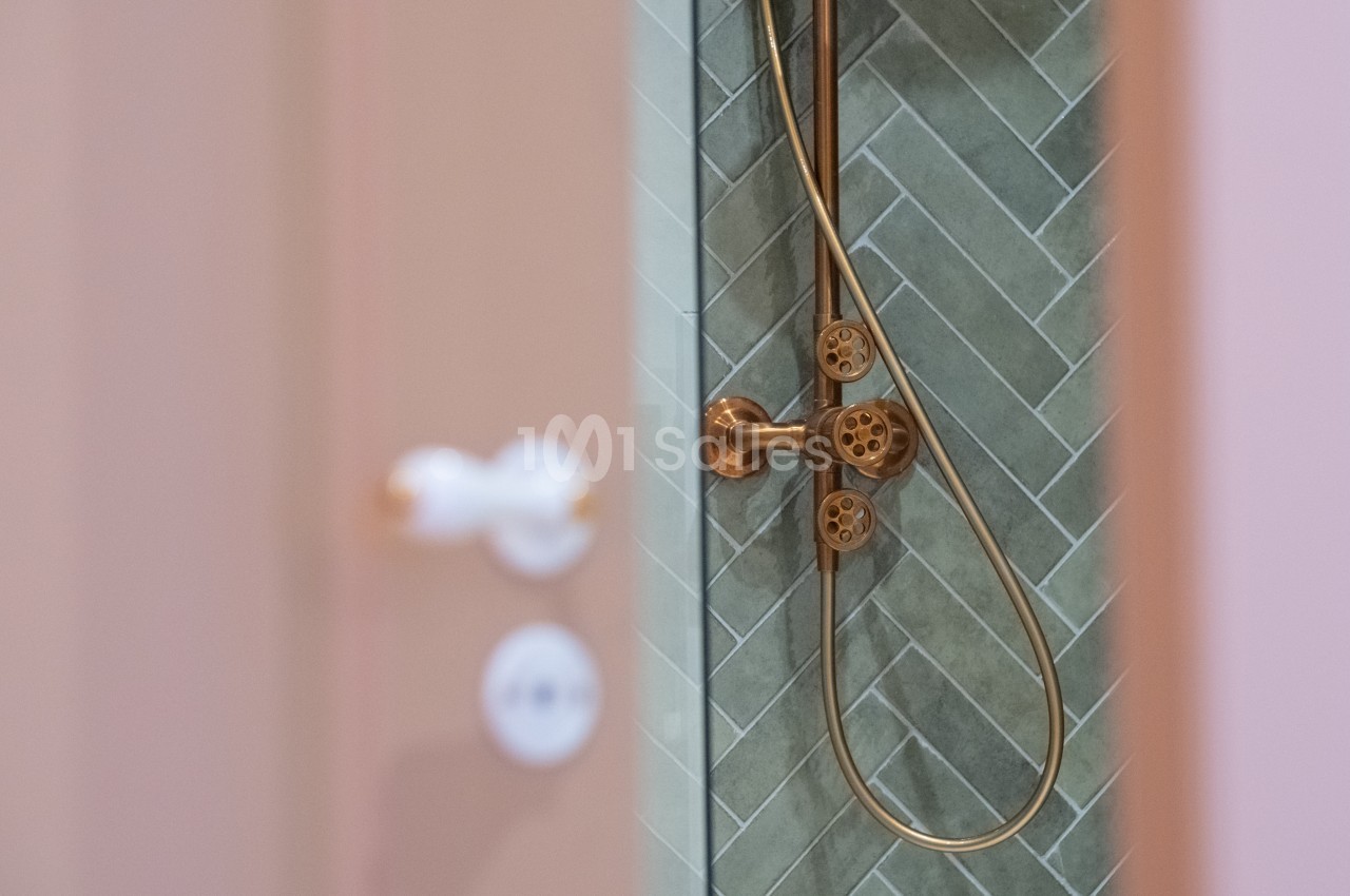 Douche avec robinetterie dorée et carrelage mural vert en motif chevron, vue partielle à travers une porte.