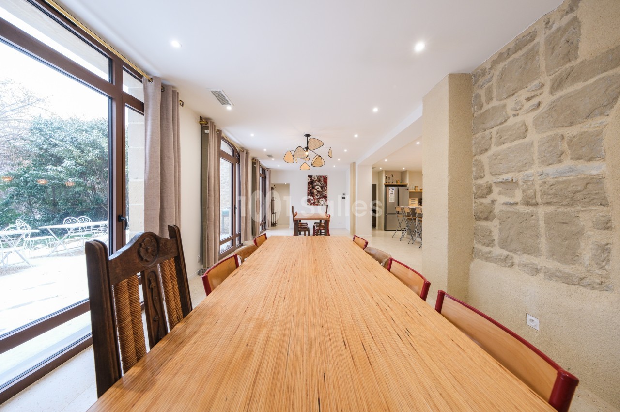 Salle à manger lumineuse avec une grande table en bois, des chaises variées et vue sur un jardin par de larges baies vitrées.