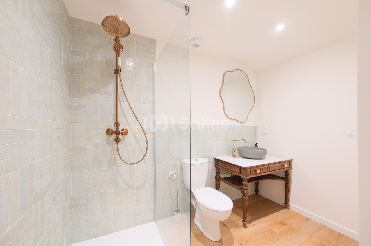 Salle de bain avec douche à l'italienne, robinetterie dorée, lavabo sur meuble en bois et miroir mural.