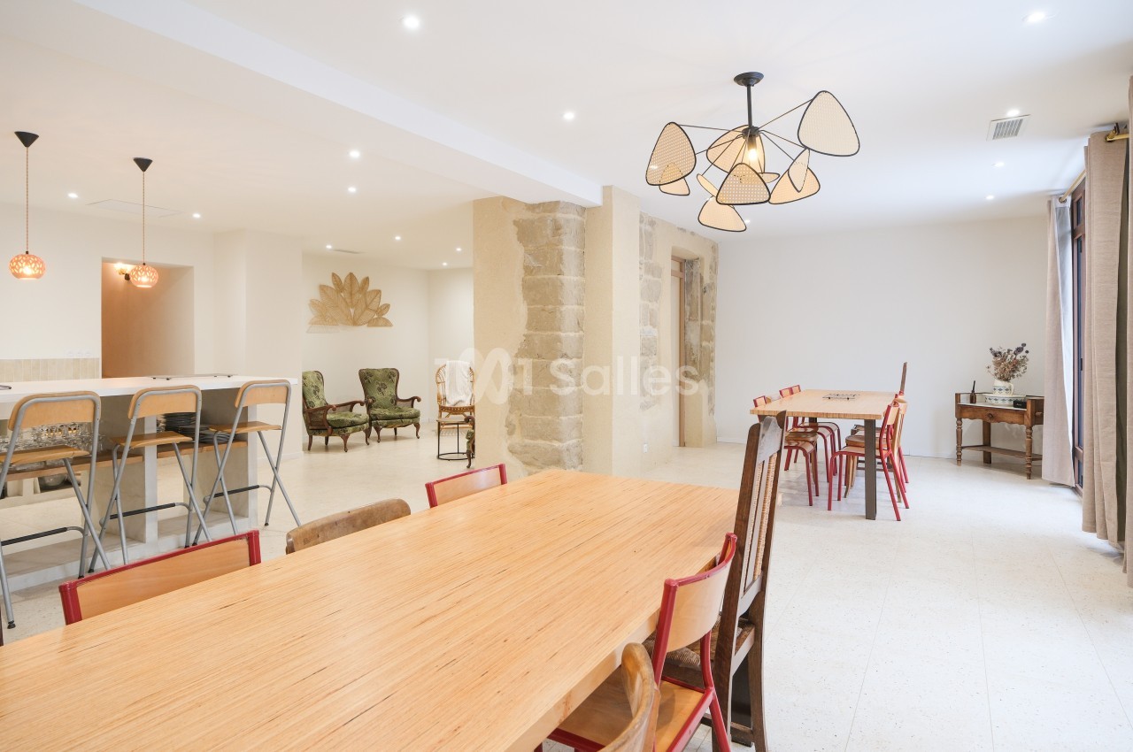 Salle lumineuse avec tables en bois, chaises variées, murs clairs, et éclairage suspendu moderne.