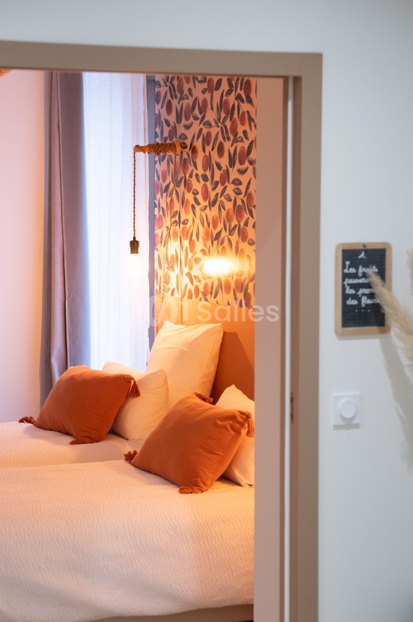 Deux lits avec coussins orange dans une chambre lumineuse, décorée de motifs floraux et éclairée par une lampe suspendue.