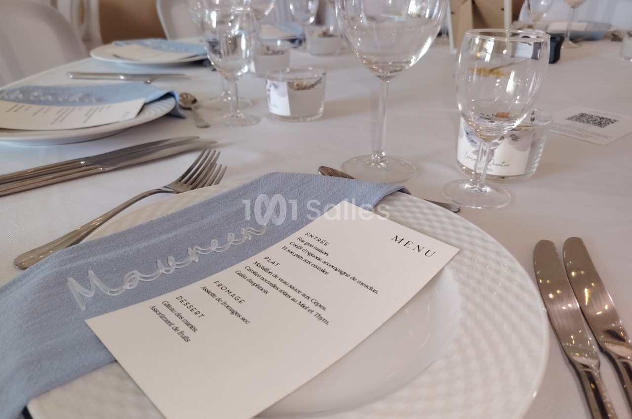 Table dressée avec une assiette, un menu, une serviette brodée et des verres pour un repas formel.