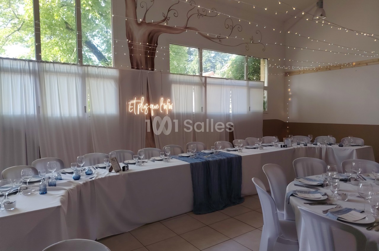 Salle de réception décorée avec des guirlandes lumineuses, une longue table dressée et un panneau lumineux ’Et ils vécurent…