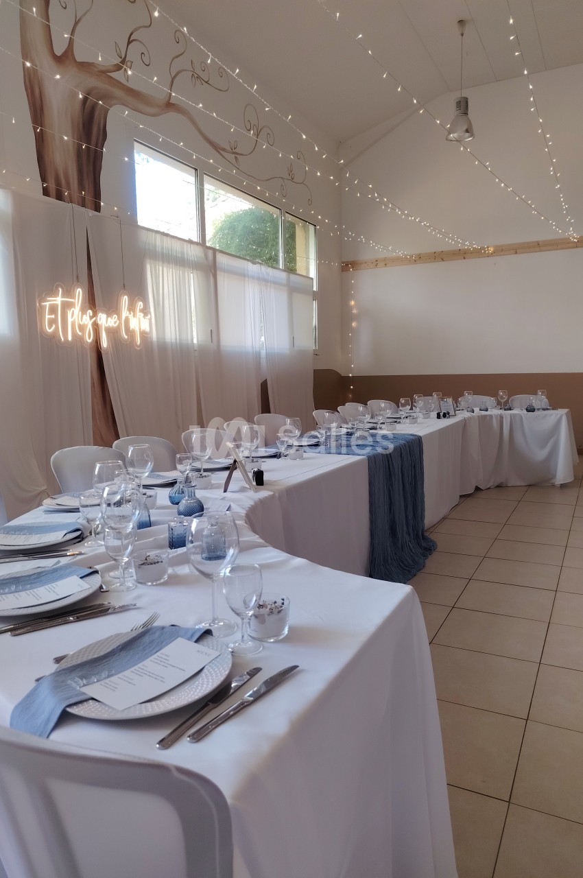 Salle décorée pour un événement avec tables dressées, nappes blanches, chemins de table bleus et guirlandes lumineuses.