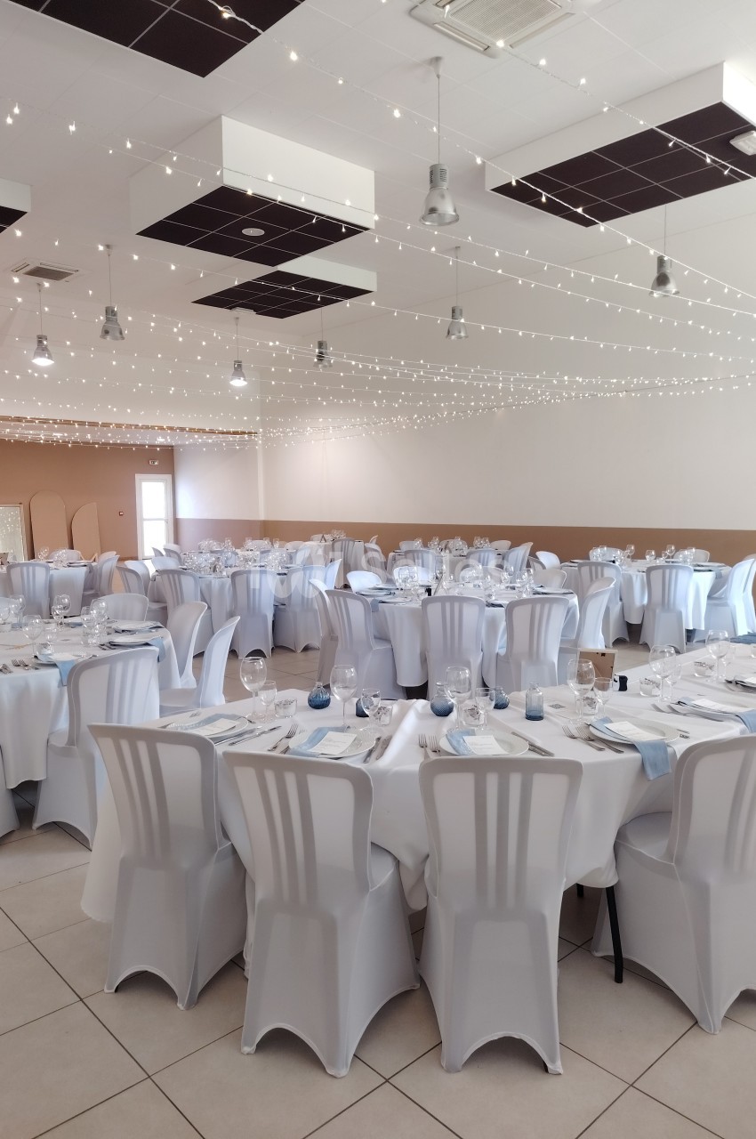 Salle de réception décorée avec des tables rondes dressées, nappes blanches et guirlandes lumineuses au plafond.