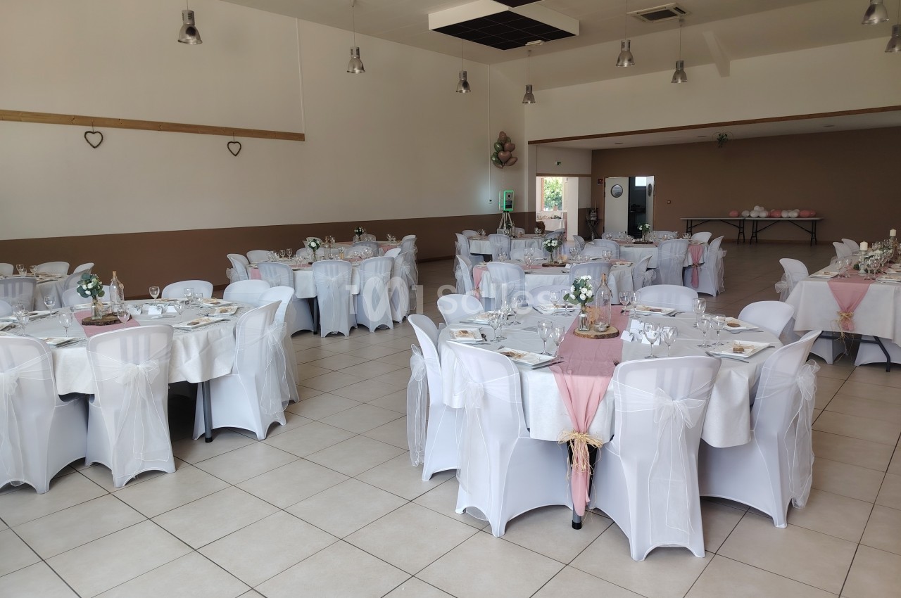 Salle de réception décorée avec des tables rondes couvertes de nappes blanches et chemins de table roses.