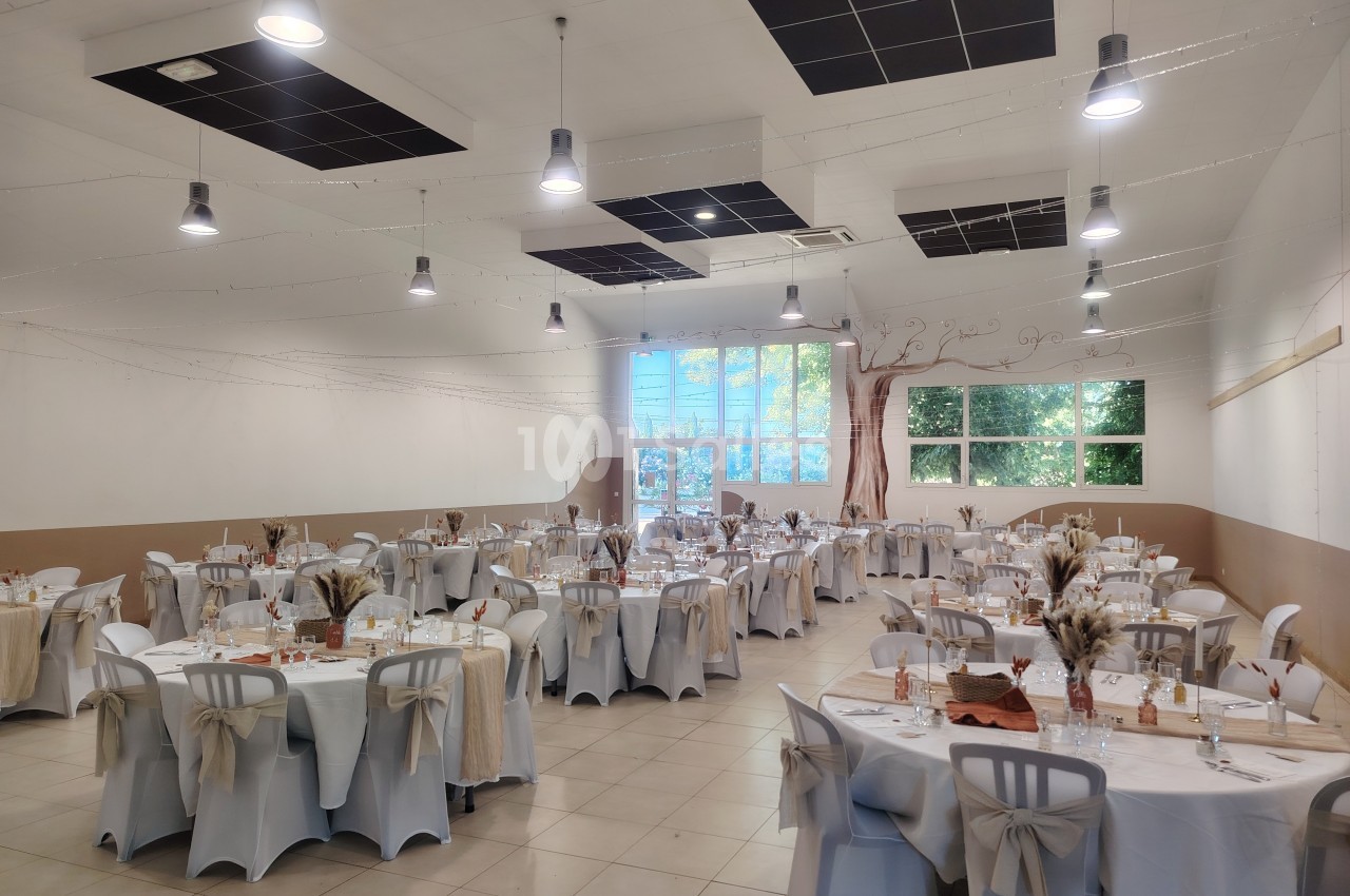 Salle de réception lumineuse décorée pour un événement, avec des tables rondes dressées et des chaises ornées de nœuds.