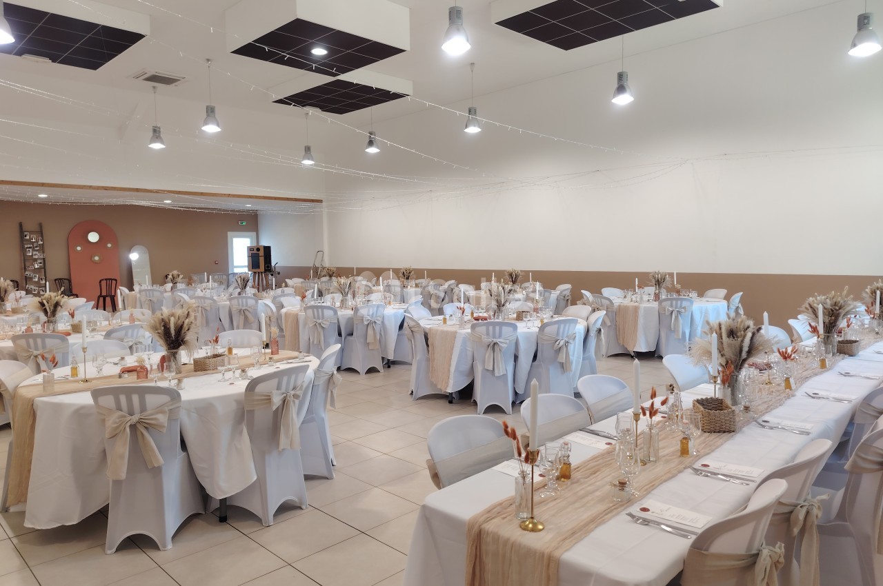 Salle de réception décorée pour un événement, avec tables dressées, nappes blanches et ornements beige et dorés.