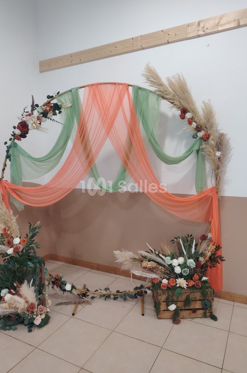 Arche décorative ornée de voilages orange et verts, fleurs séchées et feuillages, posée dans une salle claire.