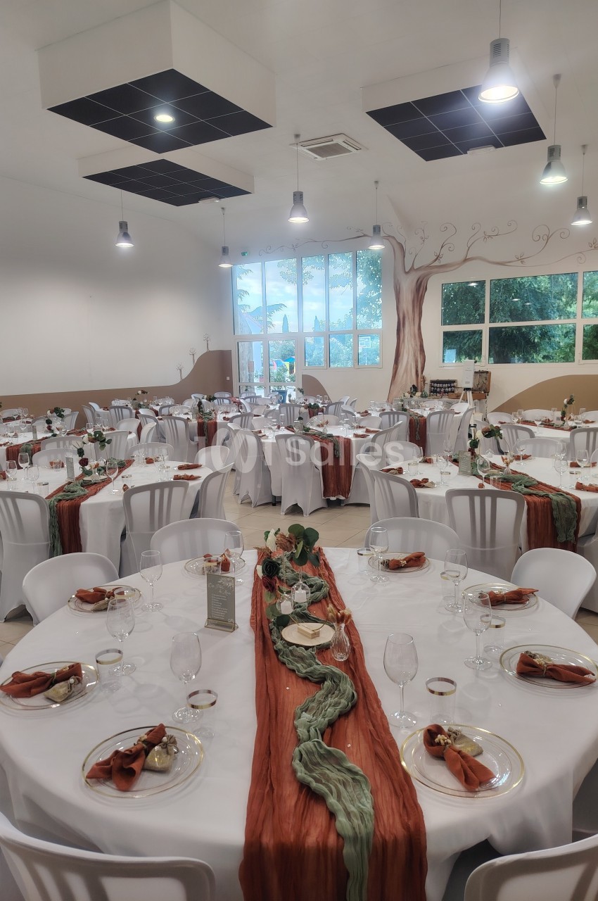 Salle de réception décorée avec des tables rondes, nappes blanches et chemins de table orange et verts.