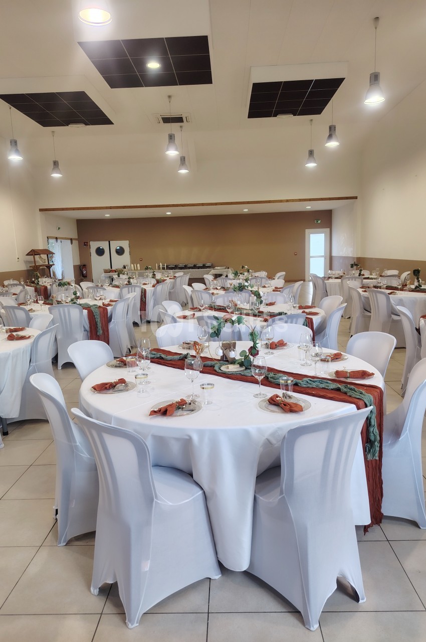 Salle de réception décorée avec des tables rondes, nappes blanches, serviettes colorées et vaisselle dressée.