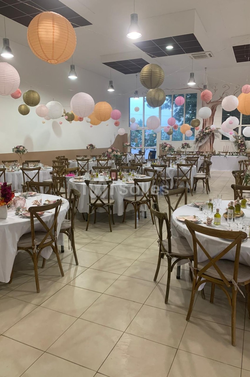 Salle décorée pour une réception avec tables rondes dressées, chaises en bois et lanternes suspendues colorées.