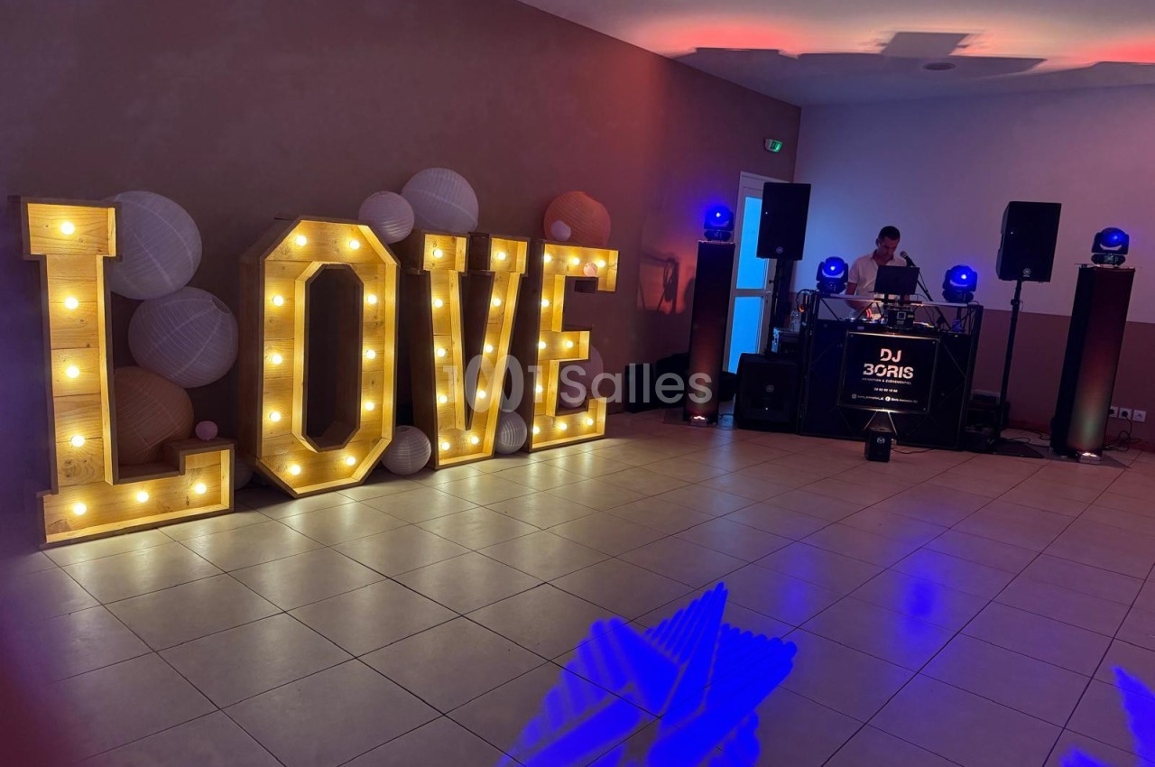 Salle de réception avec lettres lumineuses formant ’LOVE’ et un DJ en arrière-plan devant son équipement.