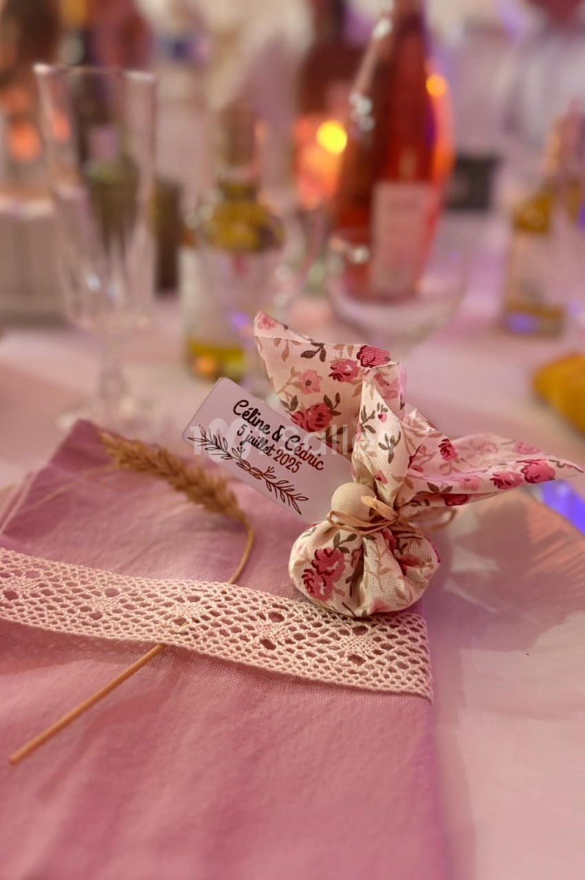 Serviette rose ornée de dentelle, épi de blé et sachet fleuri avec étiquette marquée ’Céline & Cédric, juin 2023’.