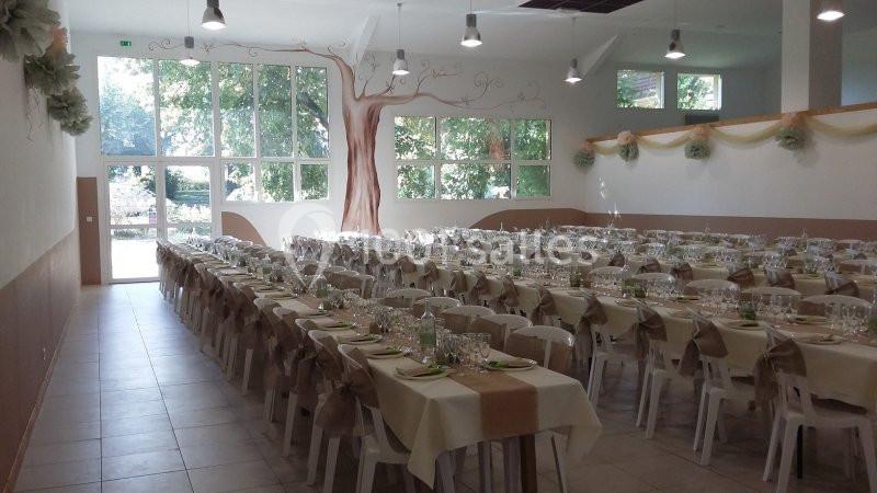 Salle de réception décorée, tables alignées avec nappes beige et blanc, couverts dressés, grandes fenêtres donnant sur des…