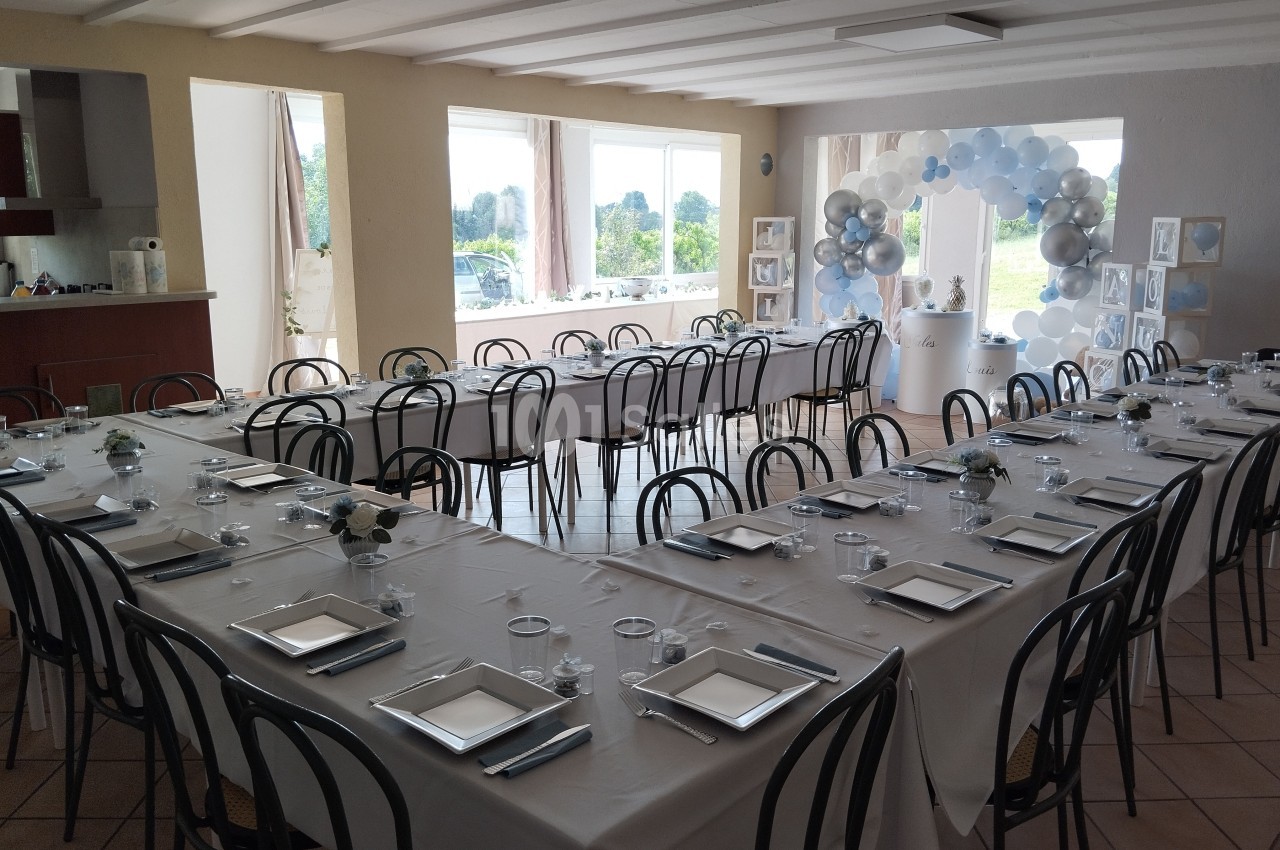 Salle décorée pour un événement, avec des tables en U dressées, des assiettes carrées et des ballons en arrière-plan.