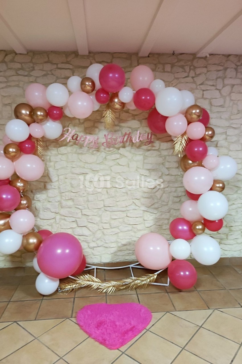 Arc de ballons roses, blancs et dorés avec inscription ’Happy Birthday’, posé devant un mur en pierre beige.