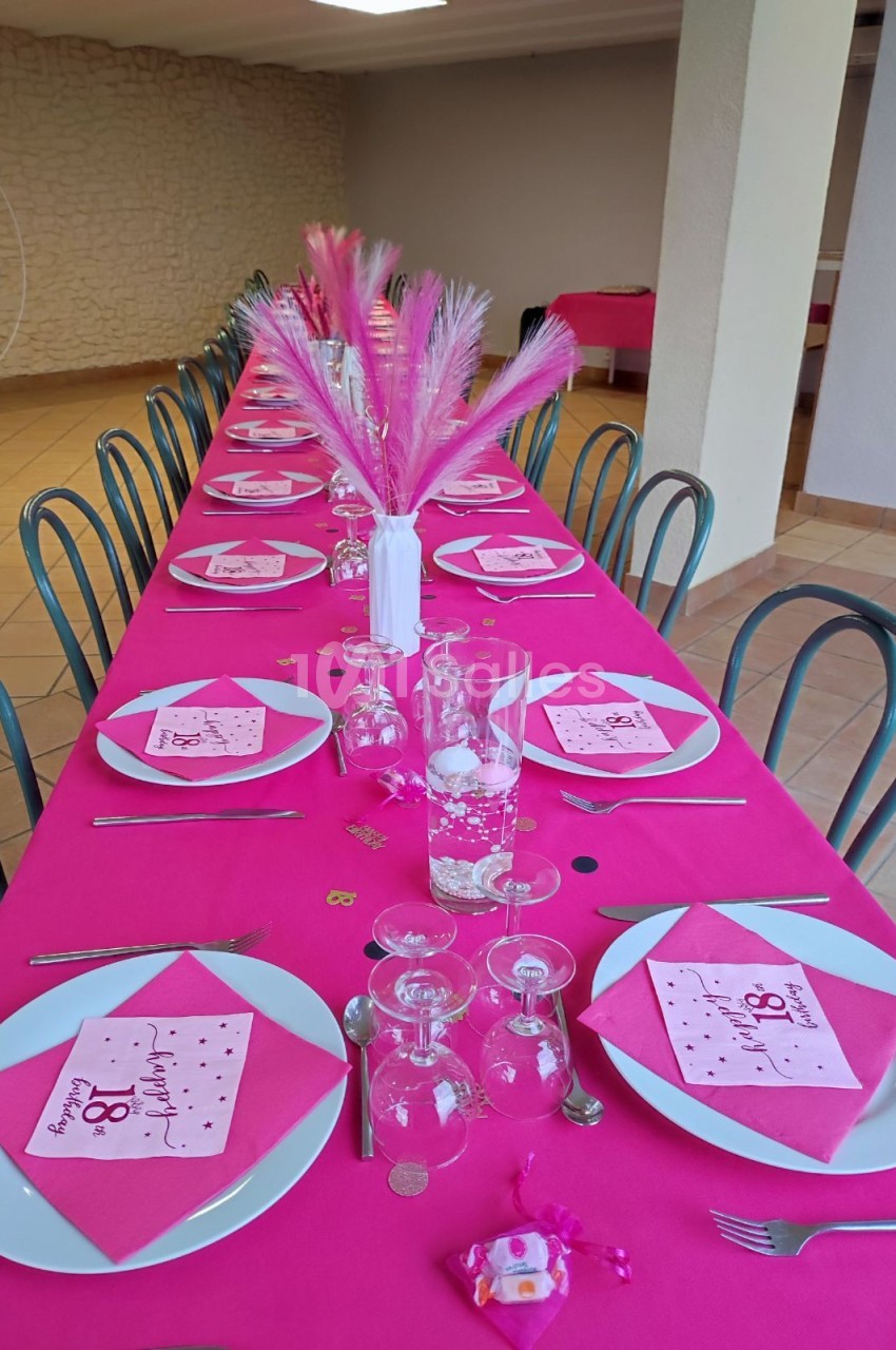 Table dressée pour une fête, nappes roses, serviettes pliées, vaisselle disposée et décoration avec plumes roses.