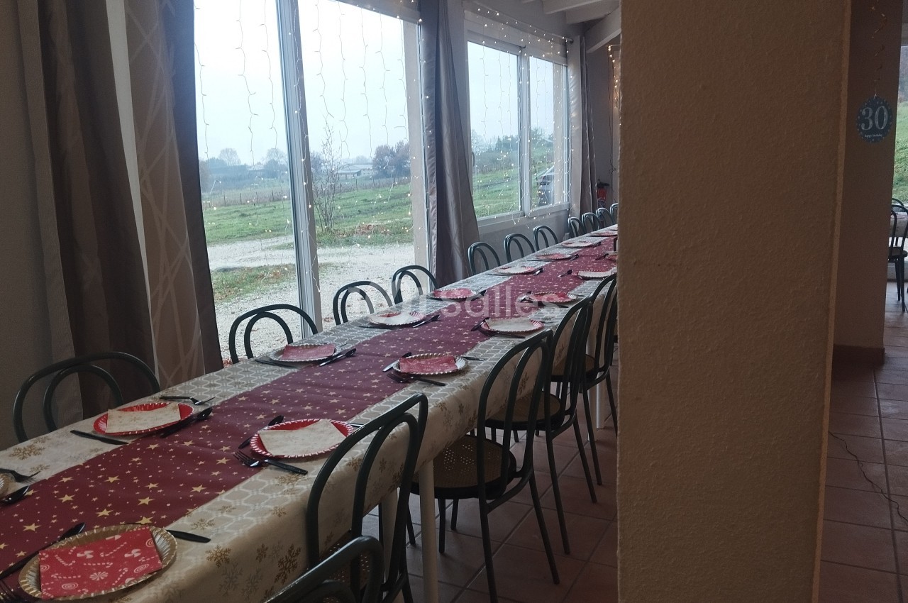 Grande table rectangulaire dressée avec nappes festives, assiettes et chaises, près de baies vitrées donnant sur un paysage…