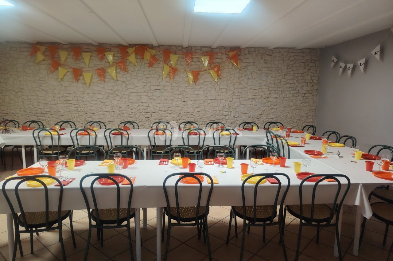 Salle décorée pour un événement, avec des tables dressées, nappes blanches, vaisselle colorée et guirlandes en fond.