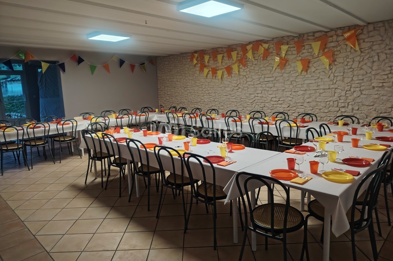 Salle décorée avec des guirlandes, tables dressées avec nappes blanches, assiettes et gobelets colorés.