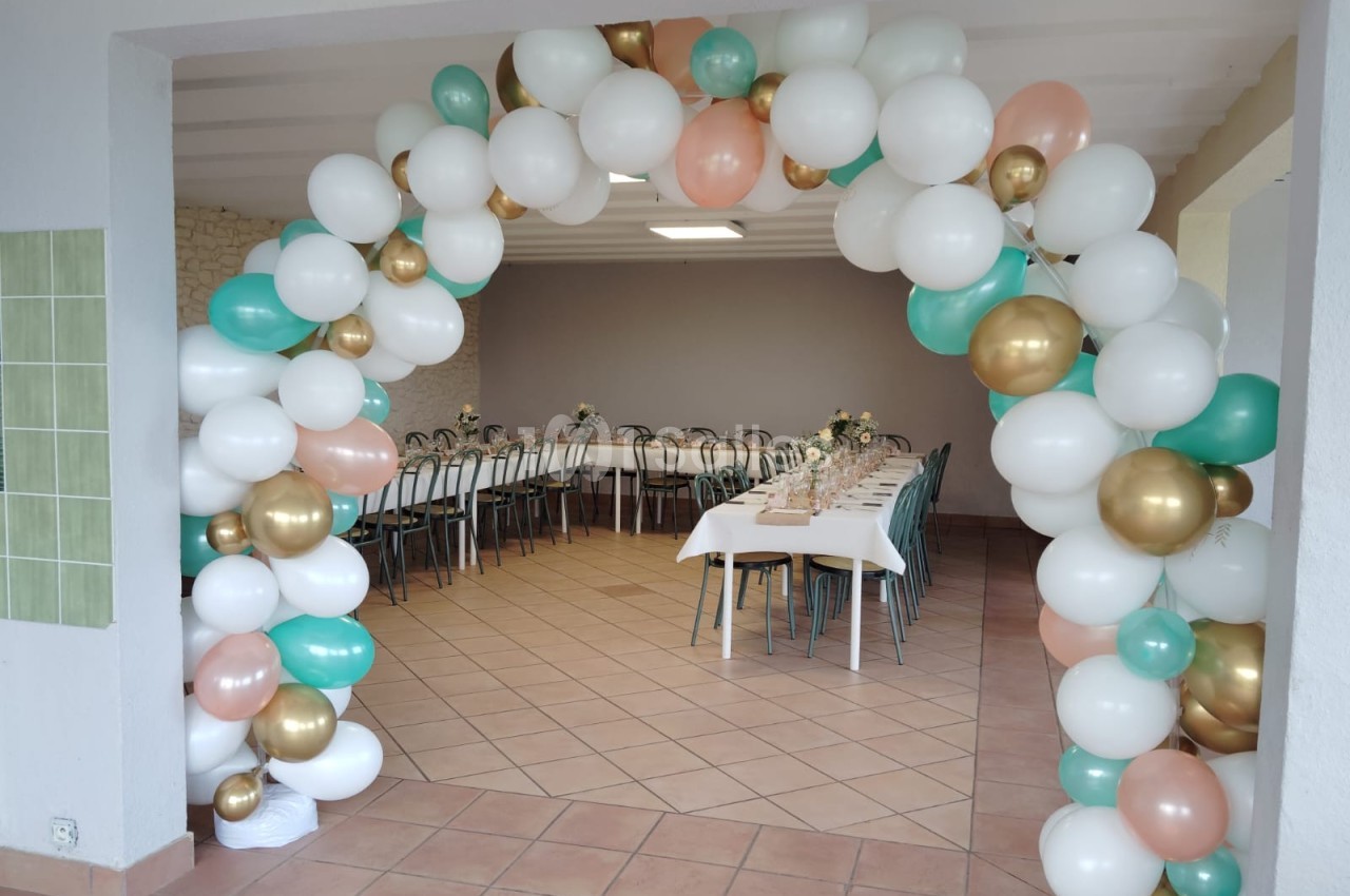 Arche de ballons blancs, verts et dorés encadrant une salle décorée avec des tables dressées pour un événement.