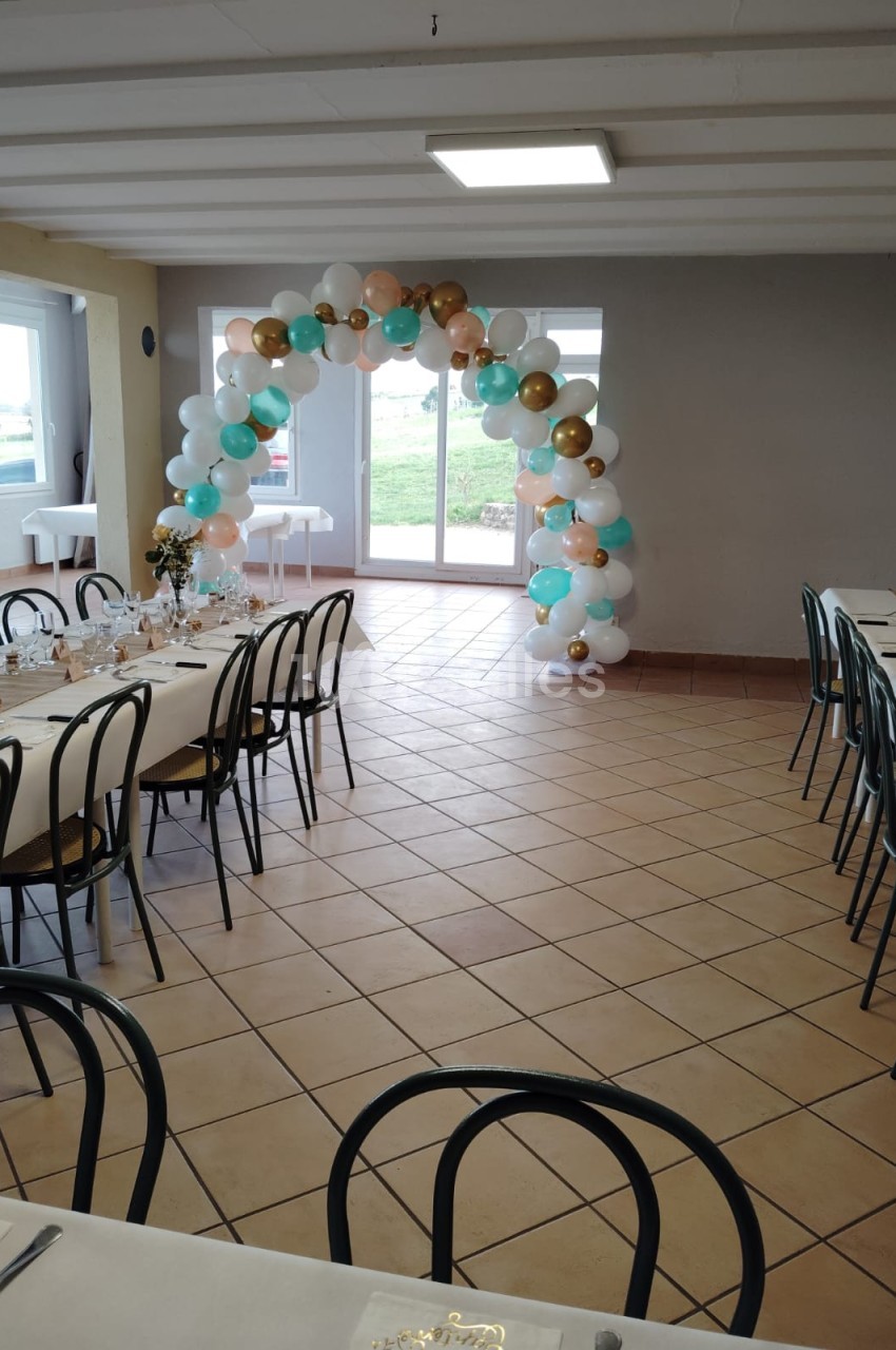 Salle décorée pour un événement avec tables dressées et arche de ballons blanc, doré et turquoise près d'une baie vitrée.