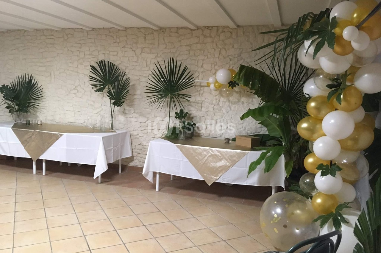 Salle décorée pour un événement avec des tables recouvertes de nappes dorées, des plantes et des ballons blancs et dorés.