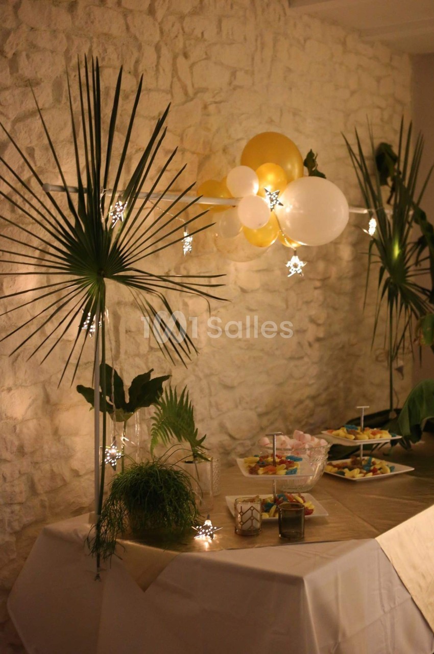 Table décorée avec des plantes, guirlandes lumineuses, ballons blancs et dorés, et plats disposés pour une réception.
