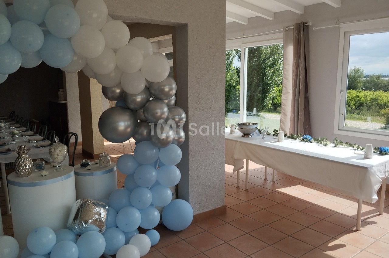 Décoration de salle avec arche de ballons bleus et blancs, table dressée et vue sur un jardin en arrière-plan.