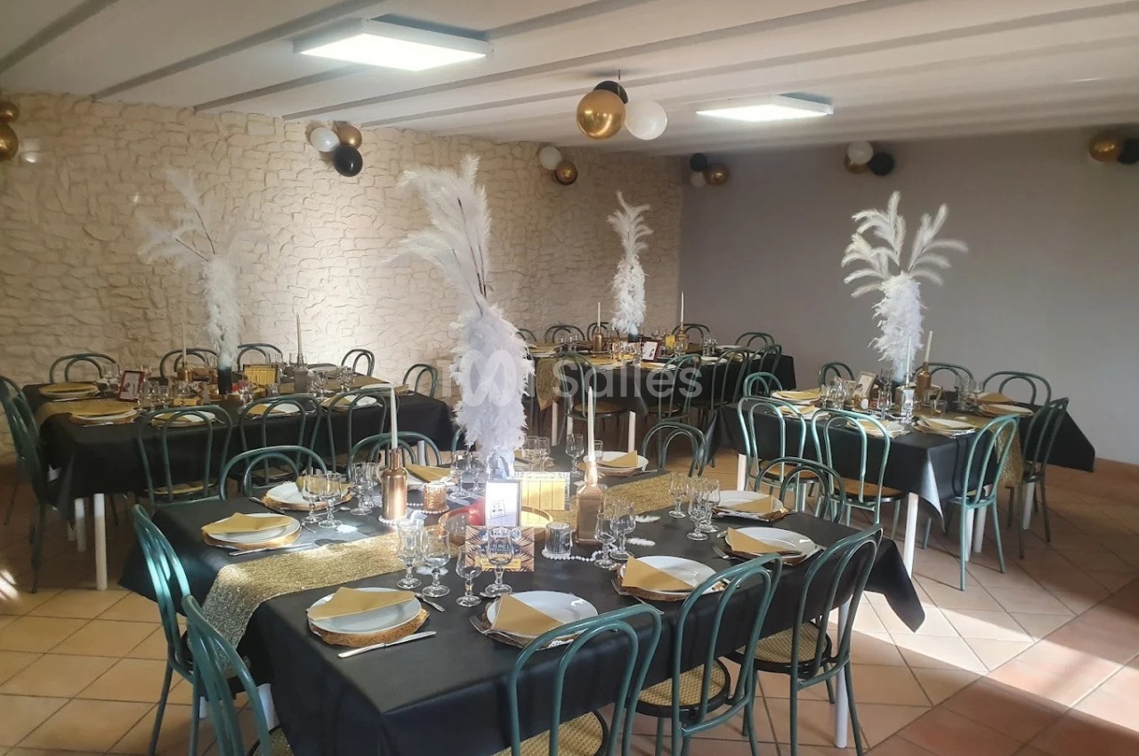 Salle décorée pour un événement, avec tables dressées, nappes noires, centres de table à plumes blanches et ballons dorés.