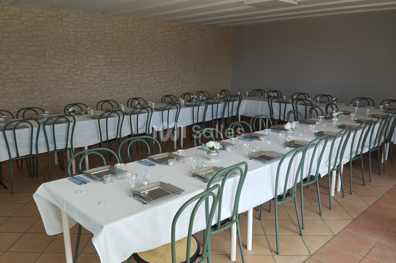 Salle aménagée avec des tables rectangulaires dressées, nappes blanches et chaises en métal, prête pour un repas.