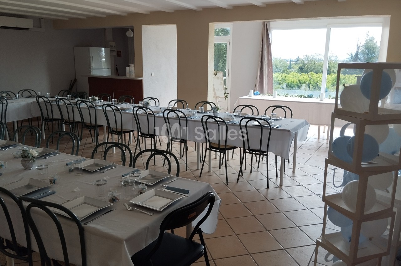 Salle lumineuse aménagée avec de longues tables dressées pour un repas, donnant sur un paysage extérieur verdoyant.