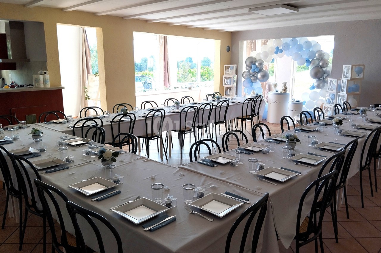 Salle décorée pour un événement avec tables dressées en U, nappes blanches et ballons en arrière-plan.
