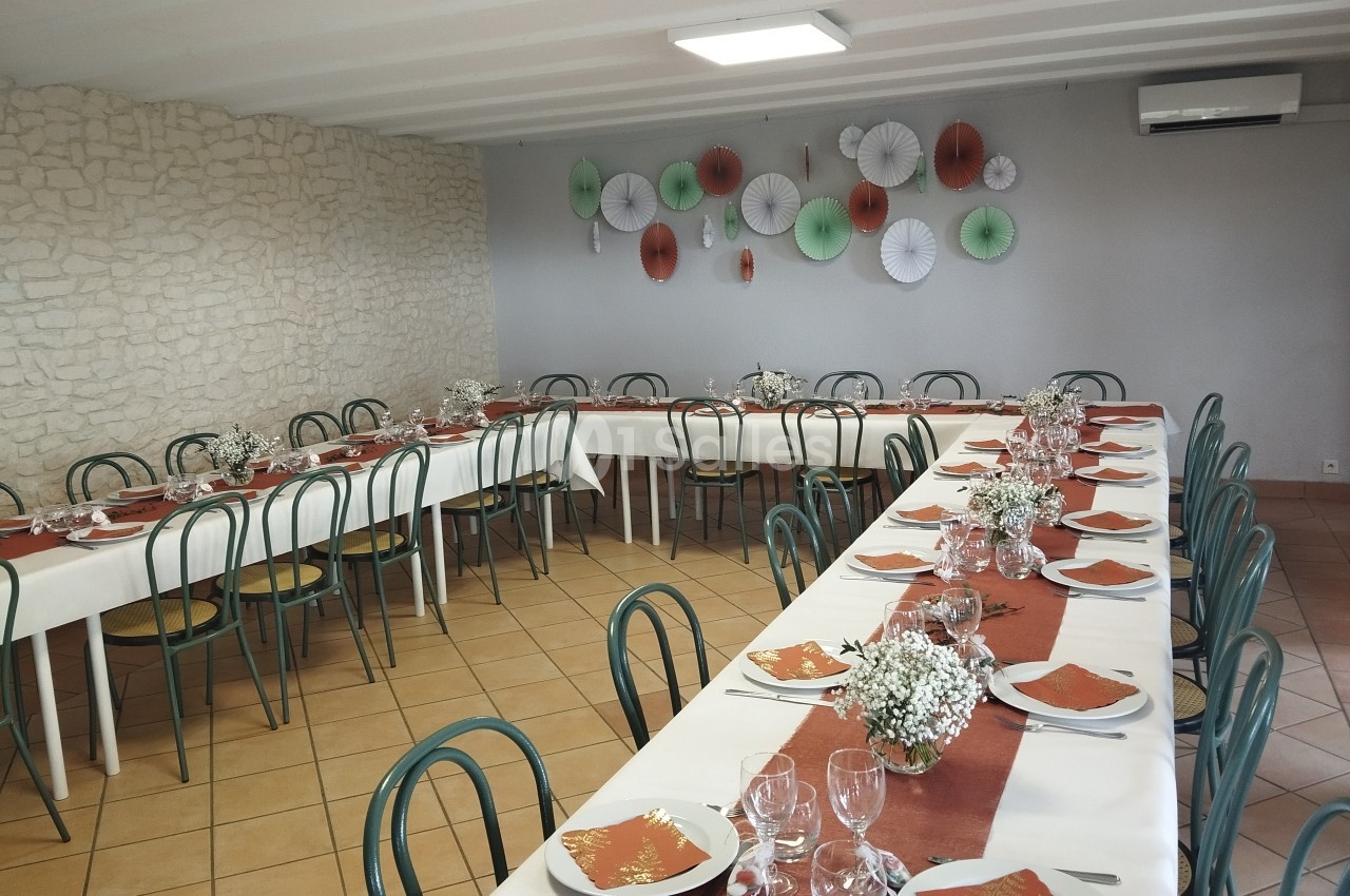Salle décorée avec des tables alignées, nappes blanches, chemins de table rouges et chaises vertes, prête pour un repas.