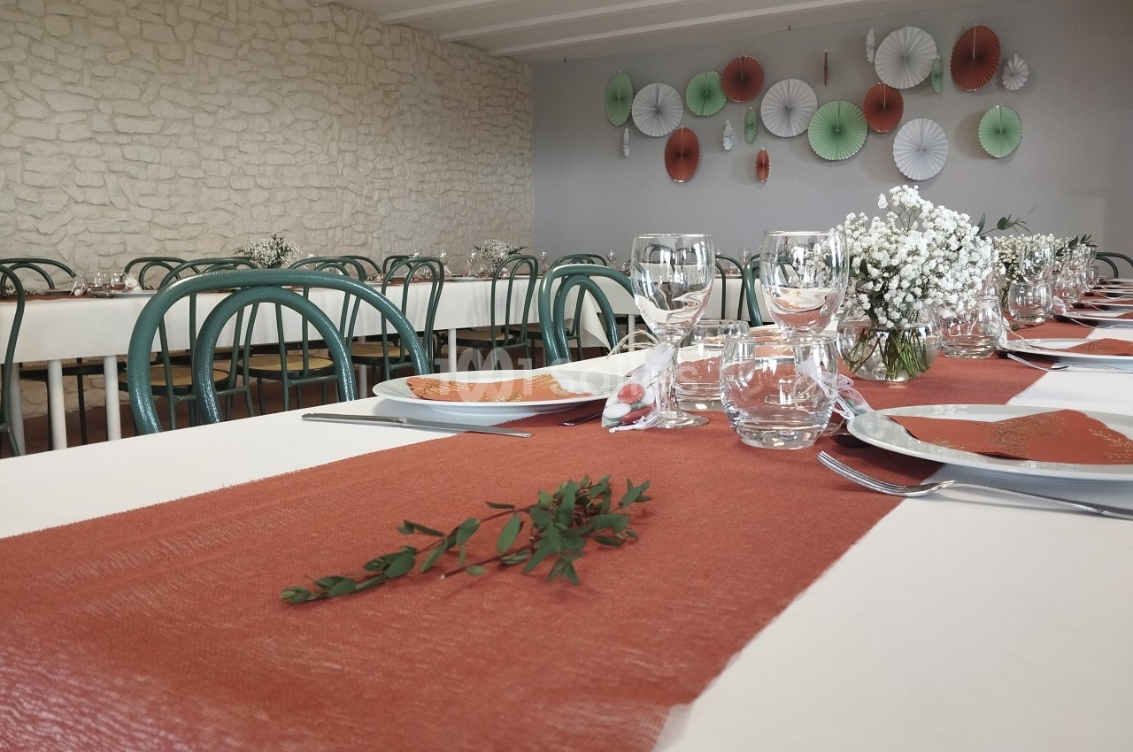 Salle décorée pour un repas, avec tables dressées, nappes blanches et chemins de table orange, ornées de fleurs.