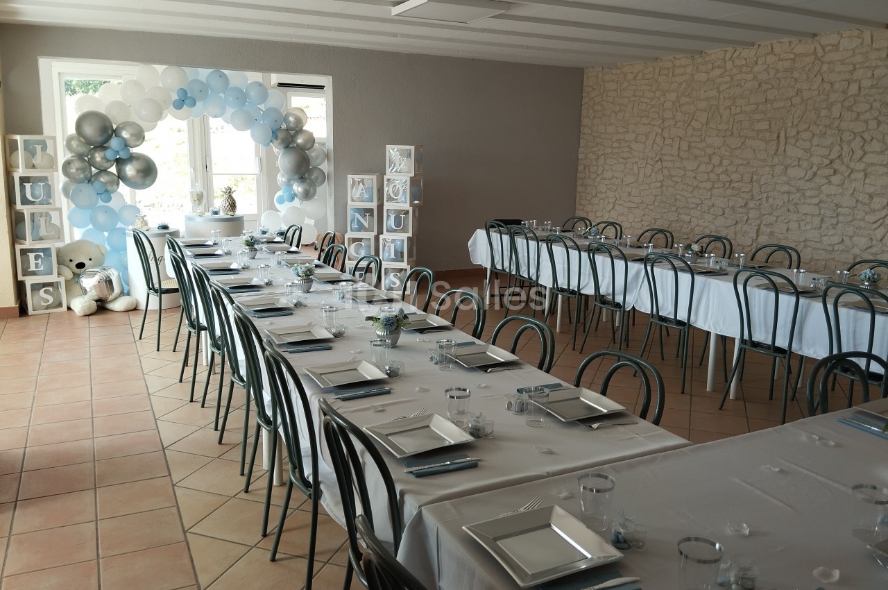 Salle décorée pour une réception avec tables dressées, ballons bleus et blancs, et cubes décoratifs près d'un ours en…