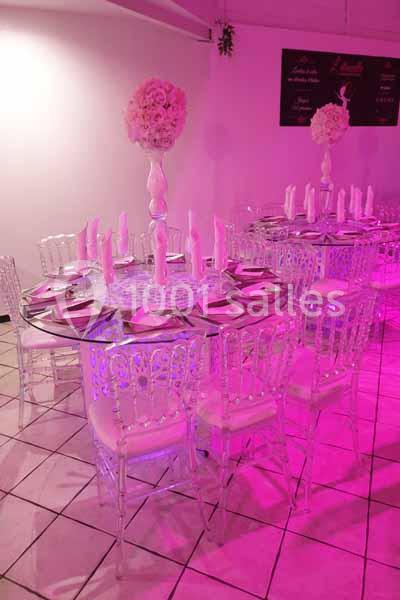 Salle décorée avec des tables rondes, nappes roses, chaises transparentes et centres de table fleuris sous éclairage rose.