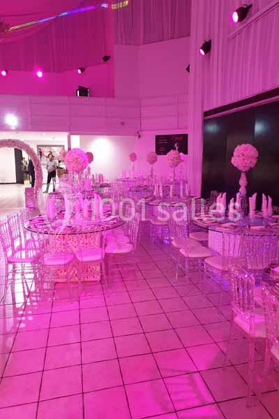 Salle de réception décorée avec des tables rondes, chaises transparentes et éclairage rose.
