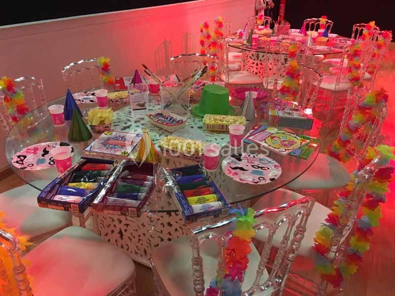 Tables rondes décorées pour une fête, avec guirlandes colorées, chapeaux, crayons et collations sous un éclairage rouge.
