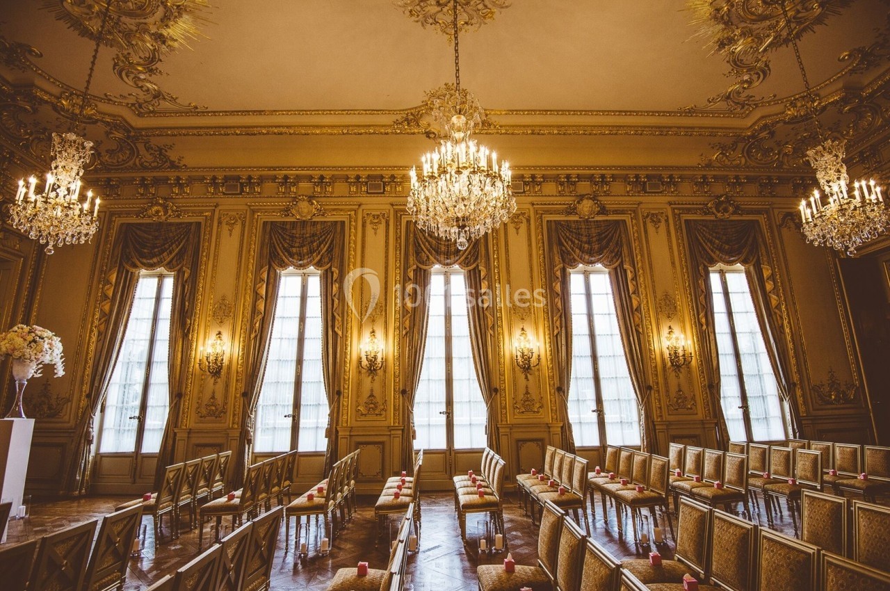 Salle de réception élégante avec lustres, hauts plafonds ornés et rangées de chaises alignées devant de grandes fenêtres.